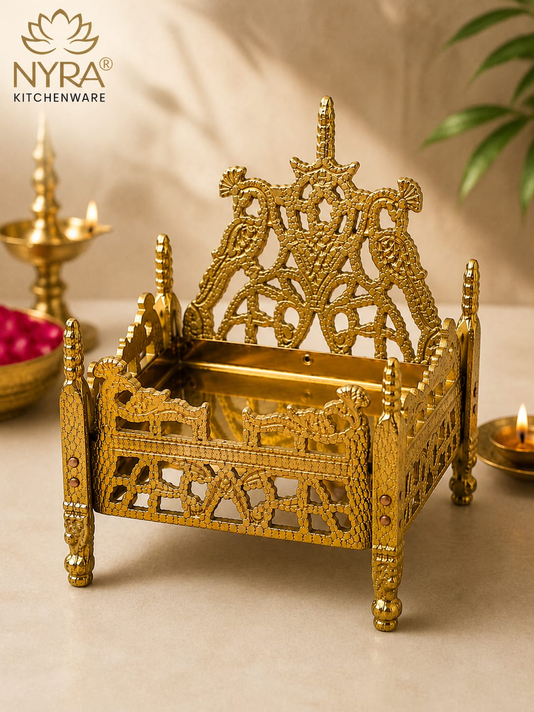 An ornate gold-plated brass miniature temple bed (Brahma Muhuttam) with intricate filigree details, displayed on a beige surface with traditional Indian diya lamps and a NYRA Kitchenware logo in the corner.