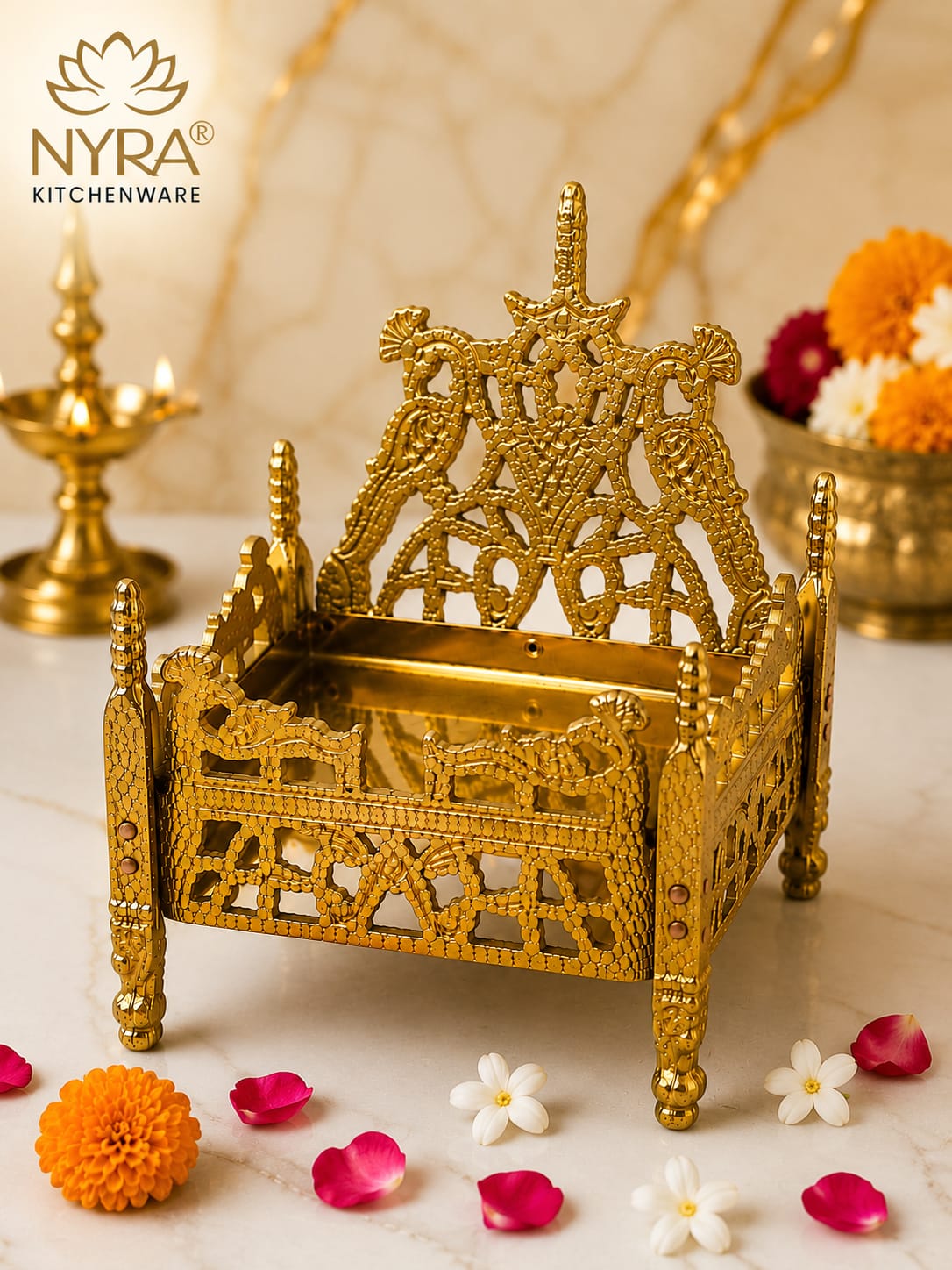 An ornate gold-plated brass miniature temple bed or decorative stand, shown on a marble surface with flowers and a diya in the background. The NYRA Kitchenware logo is visible in the top left.