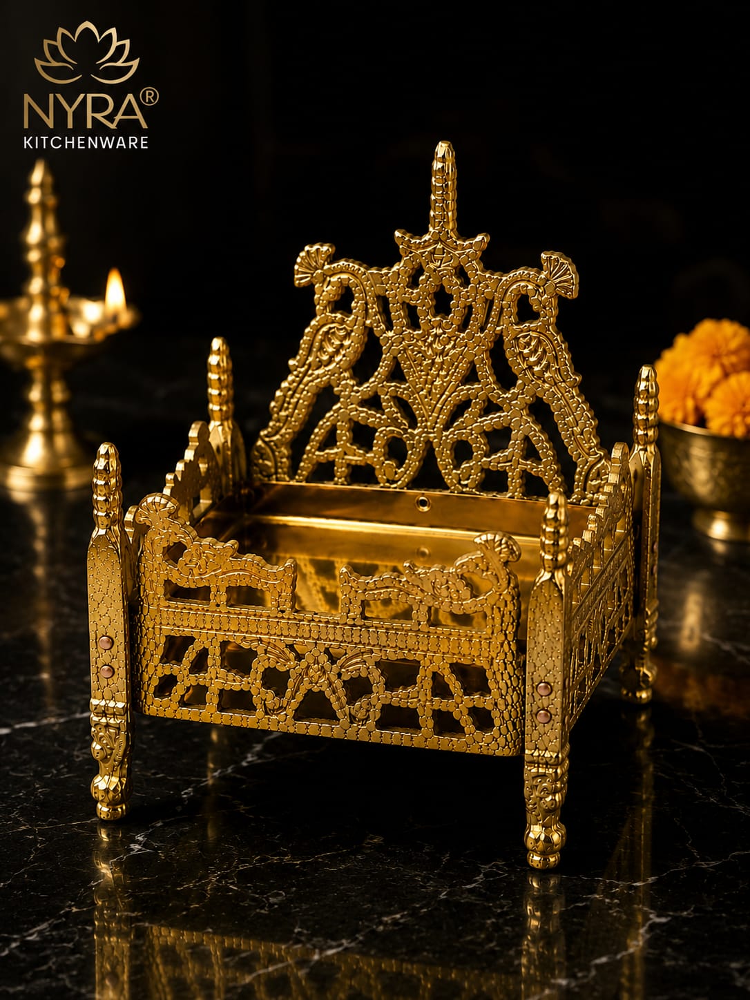 An ornate gold-plated brass Kumbhara (traditional Indian vessel) by NYRA Kitchenware, featuring intricate filigree designs and peacock motifs, displayed on a black marble surface with a lit diya in the background.