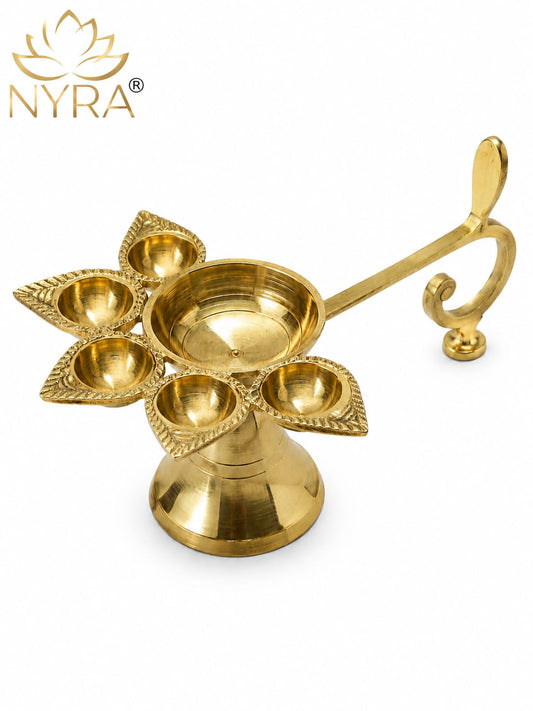 A gold brass five-light diya holder with a decorative handle, shown on a white background with the NYRA brand logo in the top left corner.