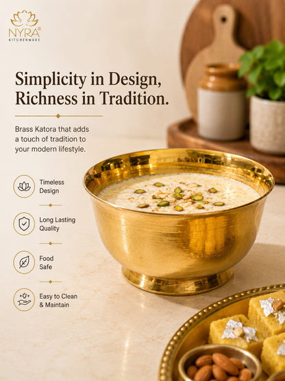 A brass Katora (traditional Indian brass garment steamer) filled with milk and topped with pistachios, presented in a polished gold-toned brass bowl on a marble surface. The background features a wooden cutting board, ceramic jars, and a plate of sweets, with the brand logo 'Nyra Kitcheness' and marketing text on the left.