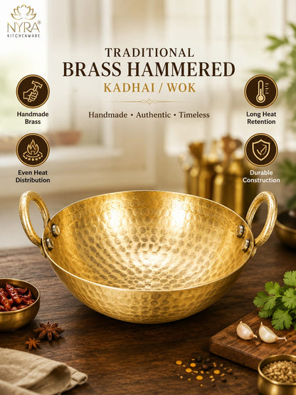 A hammered brass kadhai/wok with two side handles, shown on a wooden table with cooking ingredients and a softly blurred kitchen background. Text on the image highlights traditional brass hammered quality and listing features like handmade brass, long heat retention, and durable construction.