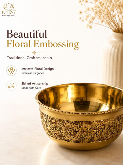 A decorative brass bowl with floral embossing on the exterior, shown on a light beige surface with a white vase of dried flowers in the background. The image includes promotional text for 'Nyra Kitchenware' and the headline 'Beautiful Floral Embossing'.
