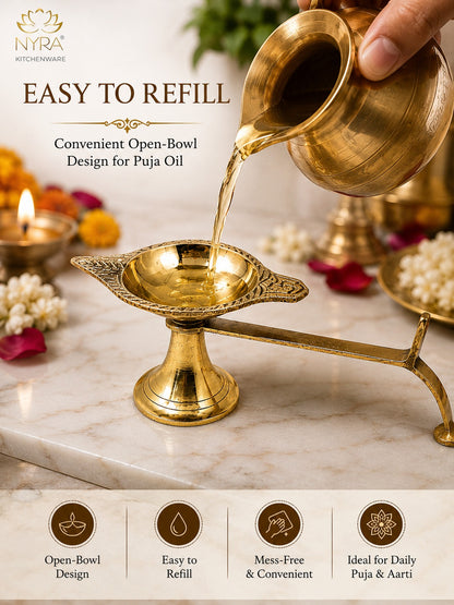 A brass puja oil lamp with a long spout and an open bowl, shown with oil being poured from a matching brass pot. The scene is styled on a marble surface with decorative flowers, a lit diya, and a hand holding the pot.