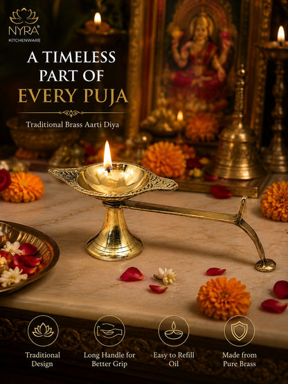 A brass Aarti diya oil lamp with a long handle, lit and set on a marble surface with marigold flowers and scattered petals, in front of a decorated puja altar. Text on the image reads 'A TIMELESS PART OF EVERY PUJA' and 'Traditional Brass Aarti Diya' with NYRA Kitchenware branding.