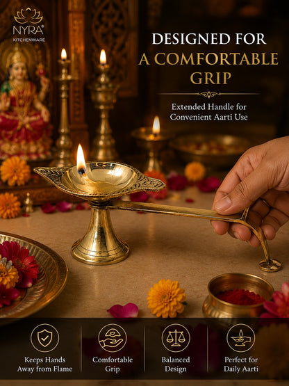 A brass oil lamp with an extended handle being held by a hand, set against a warm, decorated background with lit candles and flowers. Text on the image reads 'DESIGNED FOR A COMFORTABLE GRIP' and shows the NYRA kitchenware brand.