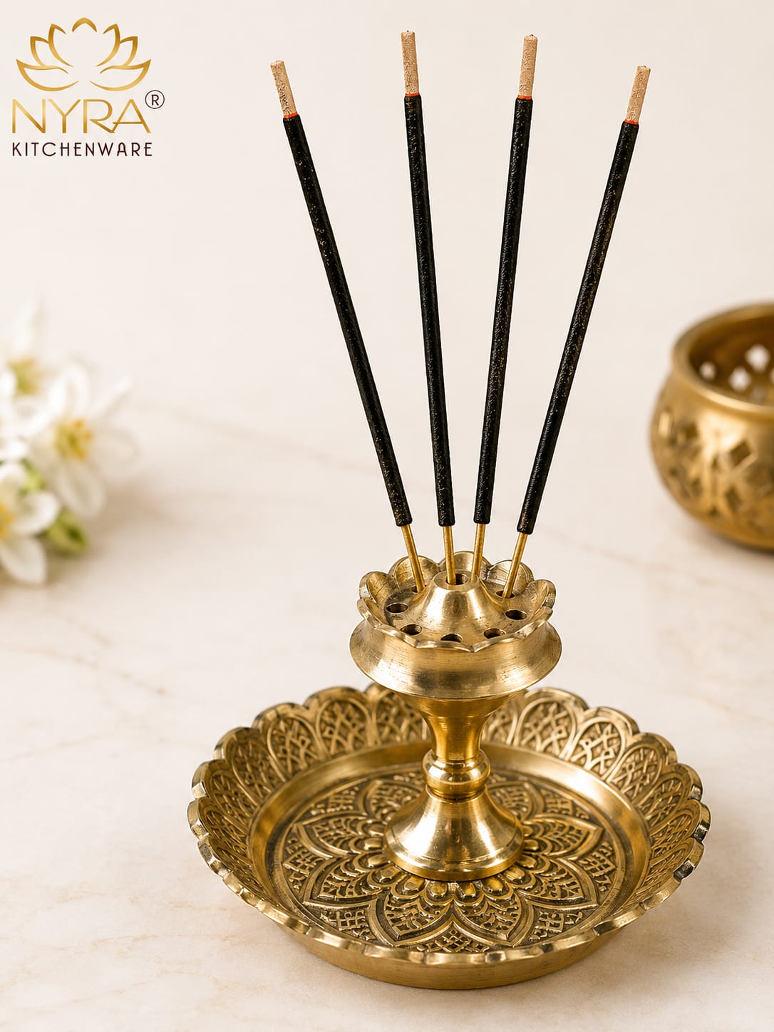 A brass incense holder with five black incense sticks, set on a light marble surface with a decorative gold-toned bowl in the background. The NYRA Kitchenware logo is visible in the top left corner.