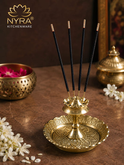 A brass incense holder with five black incense sticks, styled on a brown marble surface with white flowers and decorative brass bowls in the background. The NYRA Kitchenware logo is visible in the top left corner.