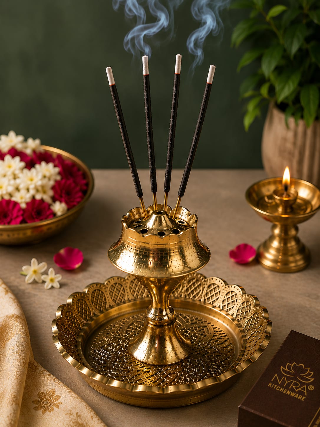 A brass incense holder with five smoking incense sticks, styled on a beige surface with a lit brass oil lamp, flowers, and a green plant in the background. A box labeled NYA KITCHENWARE is visible in the lower right corner.