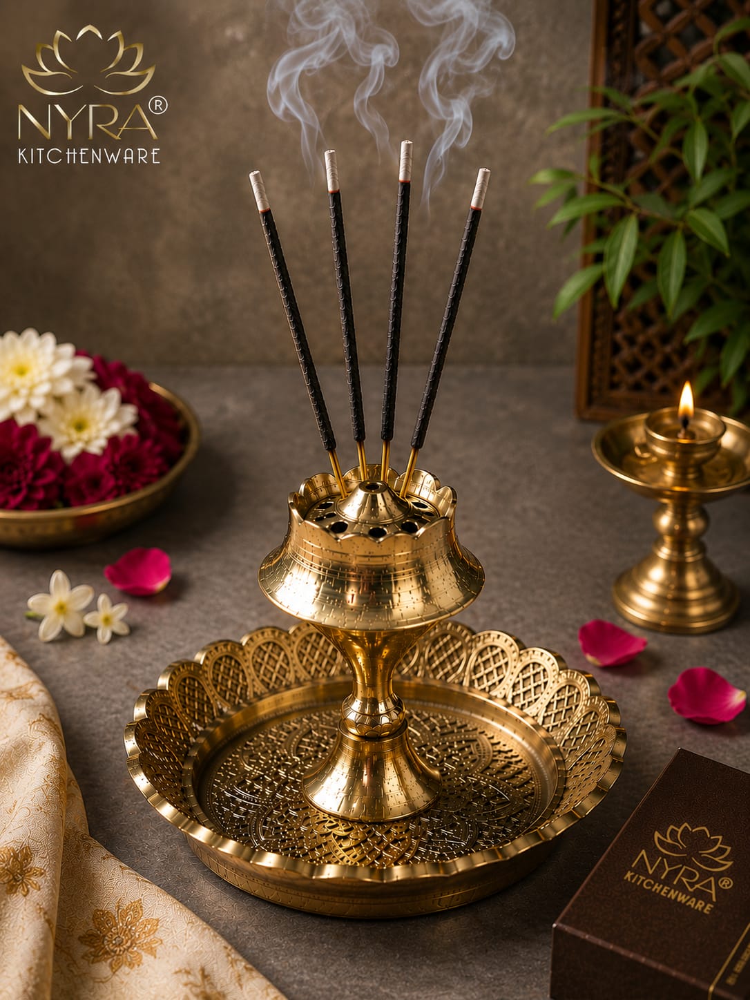 A brass double-tier incense holder with four burning incense sticks, styled on a textured gray surface with rose petals, a bowl of flowers, a small brass oil lamp, and a NYRA Kitchenware box nearby.