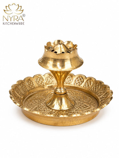 A brass double-tiered diya holder with an ornate scalloped tray and central stand, shown on a white background with the NYRA Kitchenware logo in the top left corner.