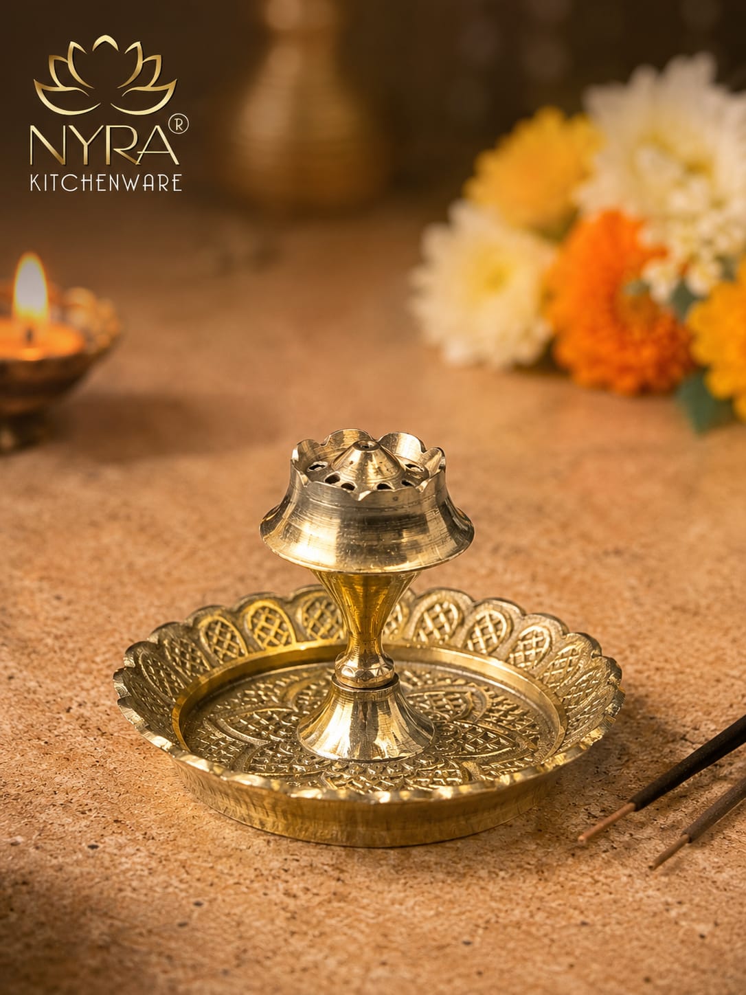 A brass oil lamp holder with an ornate scalloped tray and central lamp shade, shown on a warm beige surface with flowers and a lit candle in the background. The NYRA Kitchenware logo is visible in the top left.