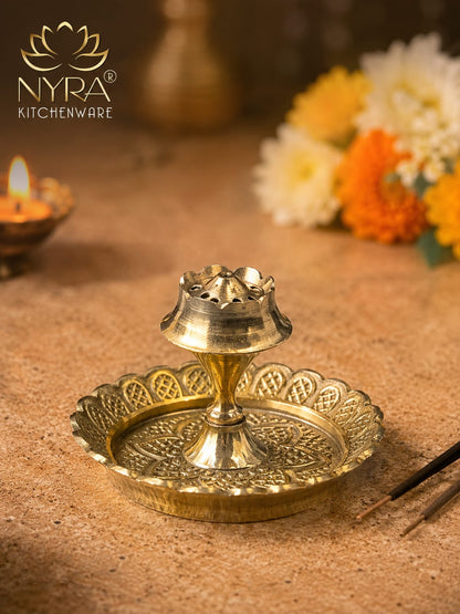 A brass oil lamp holder with an ornate scalloped tray and central lamp shade, shown on a warm beige surface with flowers and a lit candle in the background. The NYRA Kitchenware logo is visible in the top left.