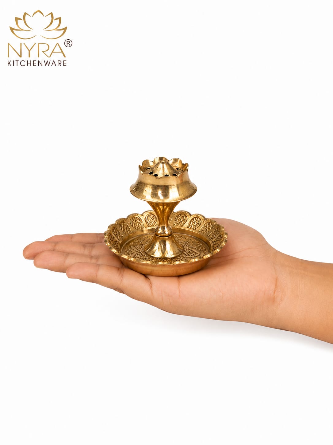 A small brass oil lamp holder with a decorative base and tiered lamp top, shown resting on an open hand against a white background. The NYRA Kitchenware logo is visible in the top-left corner.