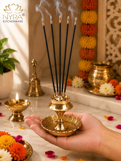 A hand holds a gold-toned brass incense holder with five smoking incense sticks. The scene is warmly lit with decorative marigold garlands, a lit oil lamp, and a small brass bell in the background, creating a rich ceremonial atmosphere.