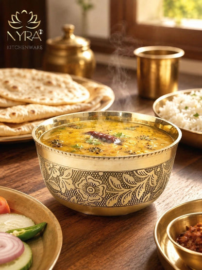 A brass bowl with floral engravings filled with a yellow curry, surrounded by Indian food on a wooden table, with the NYRA Kitchenware logo in the top left corner.