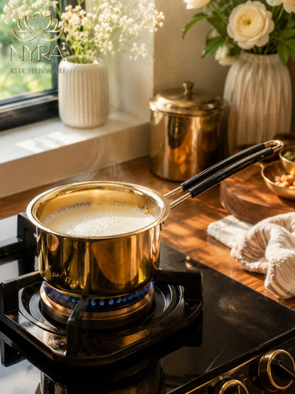 A shiny gold stainless steel saucepan with a black handle sits on a black gas stovetop, with a blue flame visible beneath. The scene is set in a warm, sunlit kitchen with flowers, a copper canister, and a cream-colored cloth on a wooden table.