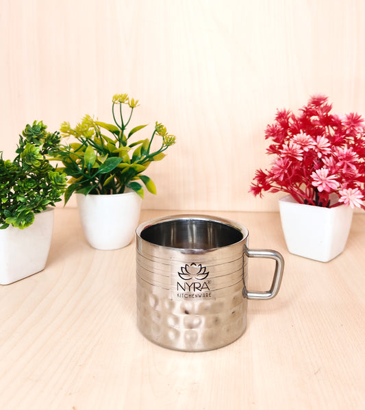 Silver mug with 'NYRA' branding on a beige surface with plants in the background