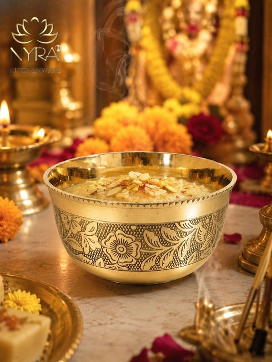 A decorative brass bowl with intricate floral engravings, filled with a warm beverage and garnished with flower petals, set on a marble surface with traditional Indian ceremonial elements in a softly blurred background. The NYRA Kitchenware logo is visible in the top left corner.